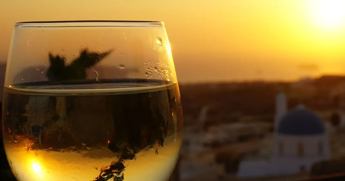 A glass of chilled Assyrtiko held against a golden Santorini sunset, a blue-domed church behind