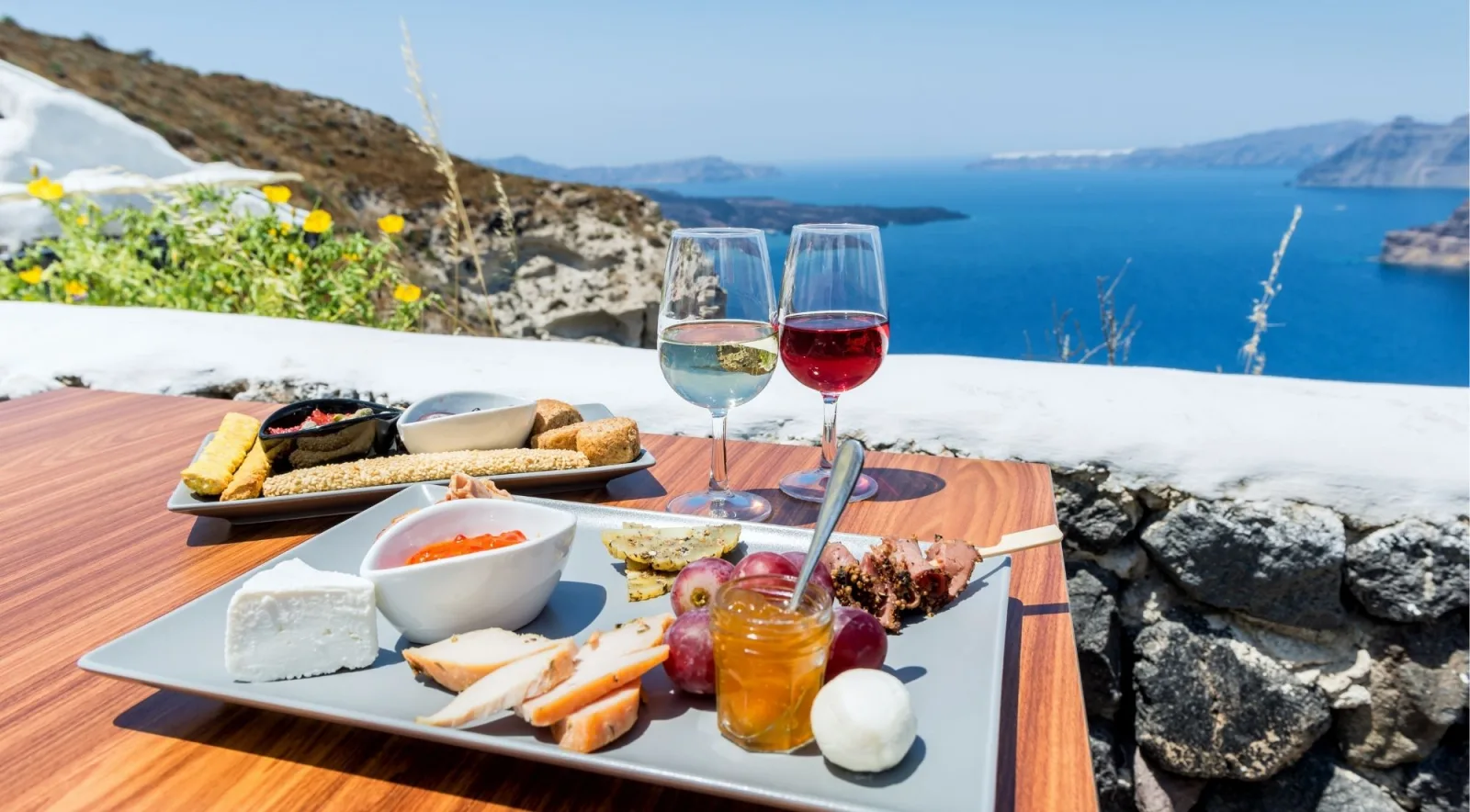 A meze platter of feta, cured meats, grapes and honey beside glasses of white and rosé wine on a terrace above the Santorini caldera