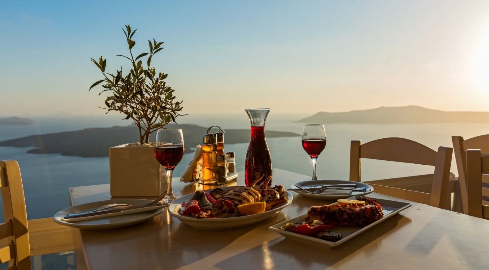 A carafe of red wine, two glasses and plates of grilled octopus on a terrace table overlooking the Santorini caldera at sunset