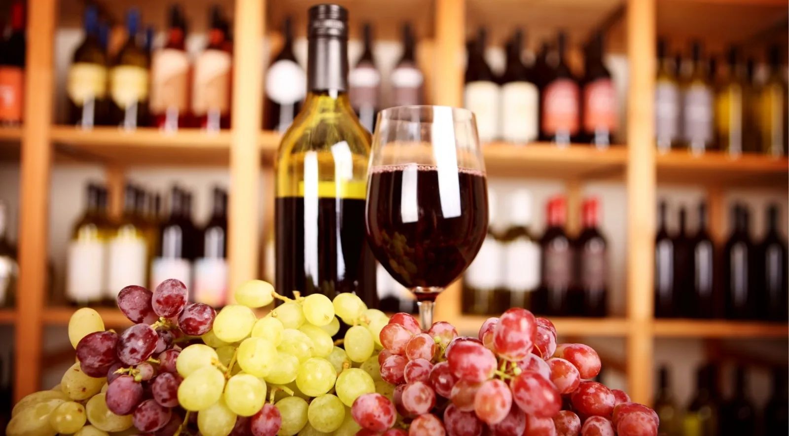 A bottle and a glass of red wine in front of shelves of wine bottles, with green and red table grapes in the foreground