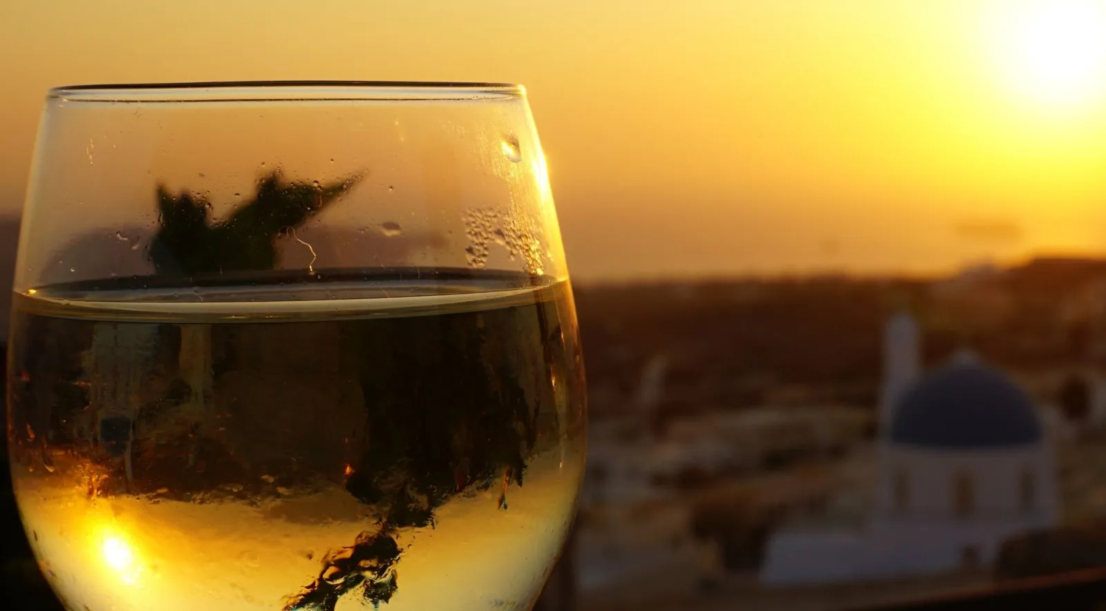 A glass of chilled white wine held against a golden Santorini sunset, with a blue-domed church blurred behind